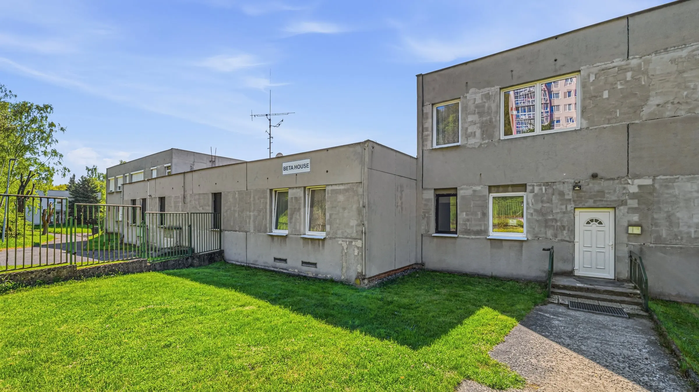 Vchod do budovy s nápisem Beta House a trávník uvnitř oploceného areálu